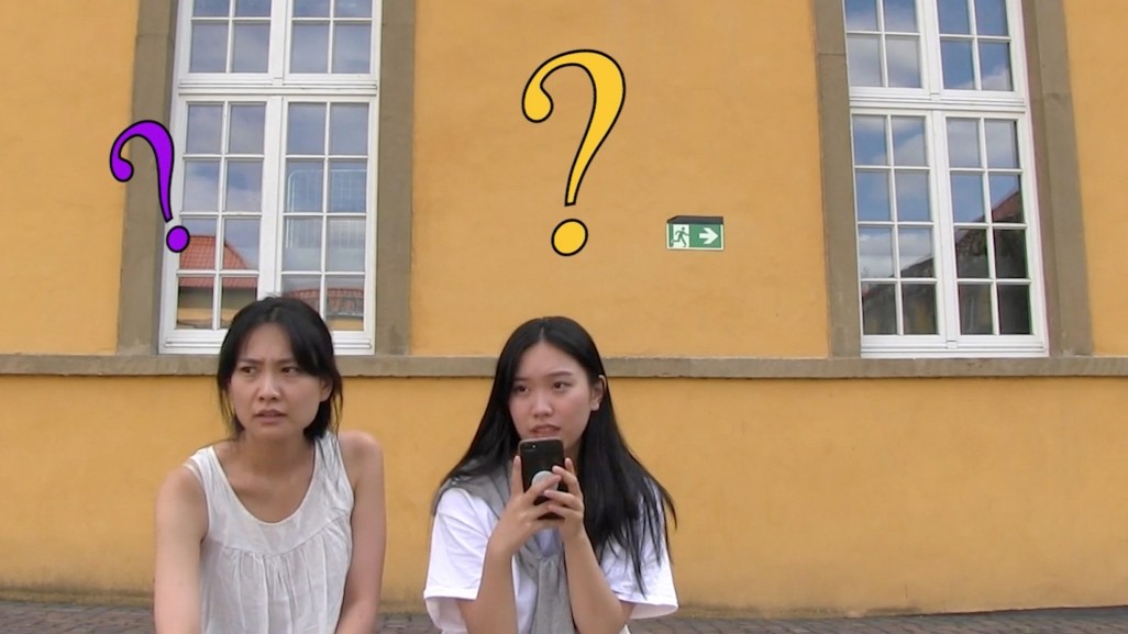 Two women sitting in front of a yellow building. Each has a question mark illustrated above her head.
